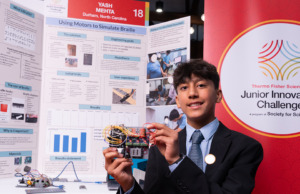 Thermo Fisher JIC finalist holds braille device in front of poster board. Finalist is wearing a black suit with a blue tie and a Thermo Fisher JIC lapel pin. 